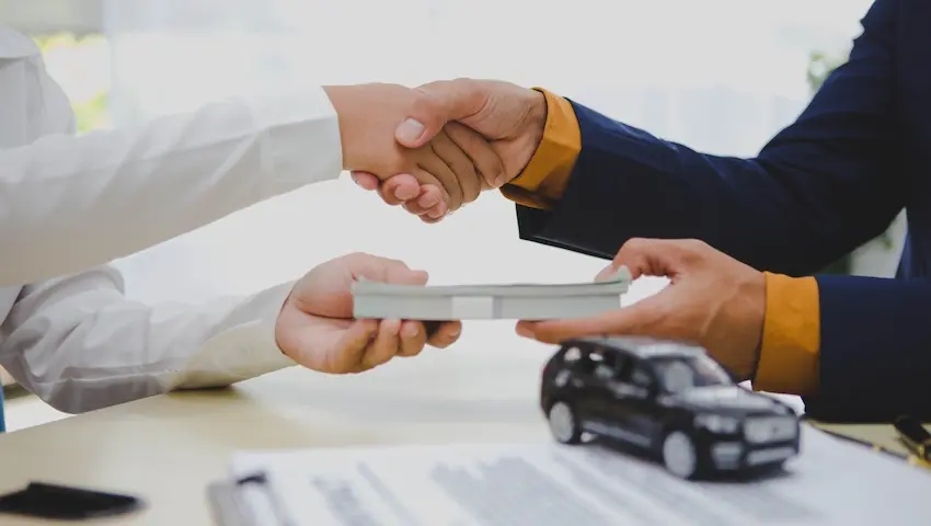 Two people shake hands across a desk while exchanging a stack of cash beside a model SUV and paperwork, representing a financial settlement that accounts for repair-related diminished value following a vehicle collision in Georgia.