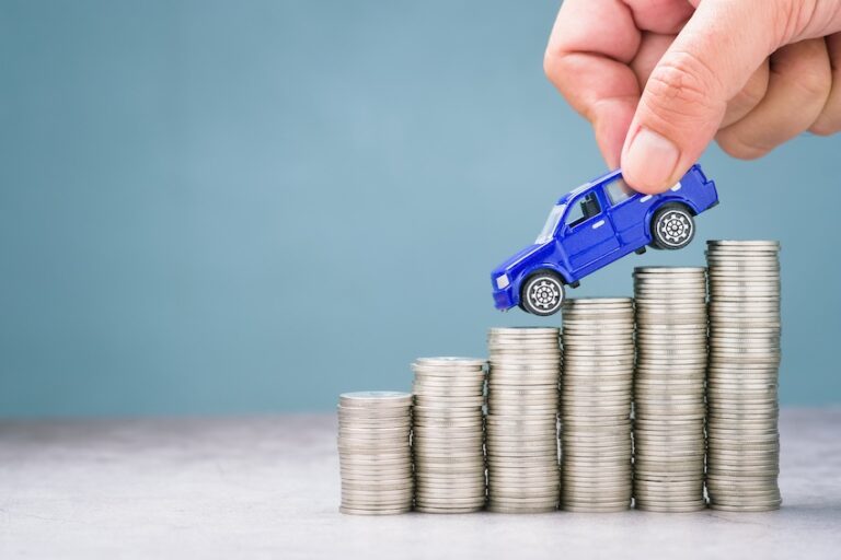 A hand places a small blue toy truck on top of ascending stacks of coins symbolizing the financial recovery process of recouping repair-related diminished value after a vehicle has been damaged and repaired with help from Claims Concierge.