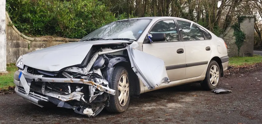 A silver sedan with severe front-end and driver-side collision damage sits parked on a road beside a stone wall, depicting the type of significant structural damage that leads to a repair-related diminished value claim in Georgia.
